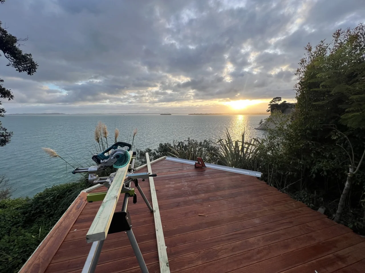 A timber deck under construction overlooking the Auckland coast at sunset, with a mitre saw and timber on site